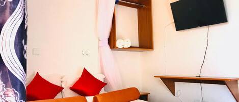 Une chambre à coucher avec un lit, des coussins rouges et orange, une table de chevet en bois, un miroir et un téléviseur fixé au mur.