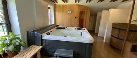 A modern spa room with a hot tub, wooden ceiling beams, a small wooden bench, and a potted plant.
