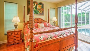 A four-poster bed with a floral bedspread, wooden nightstands with lamps, and a large window with a view of greenery. - Naples Home w/ Pool, Extended Stays Welcome! (Naples)