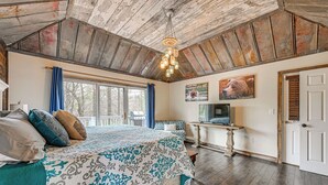 A bedroom with a large bed, a wooden ceiling, a window with curtains, a desk, and a door. - Outdoor Fireplace + Views Mountain Bearadise (Sapphire)