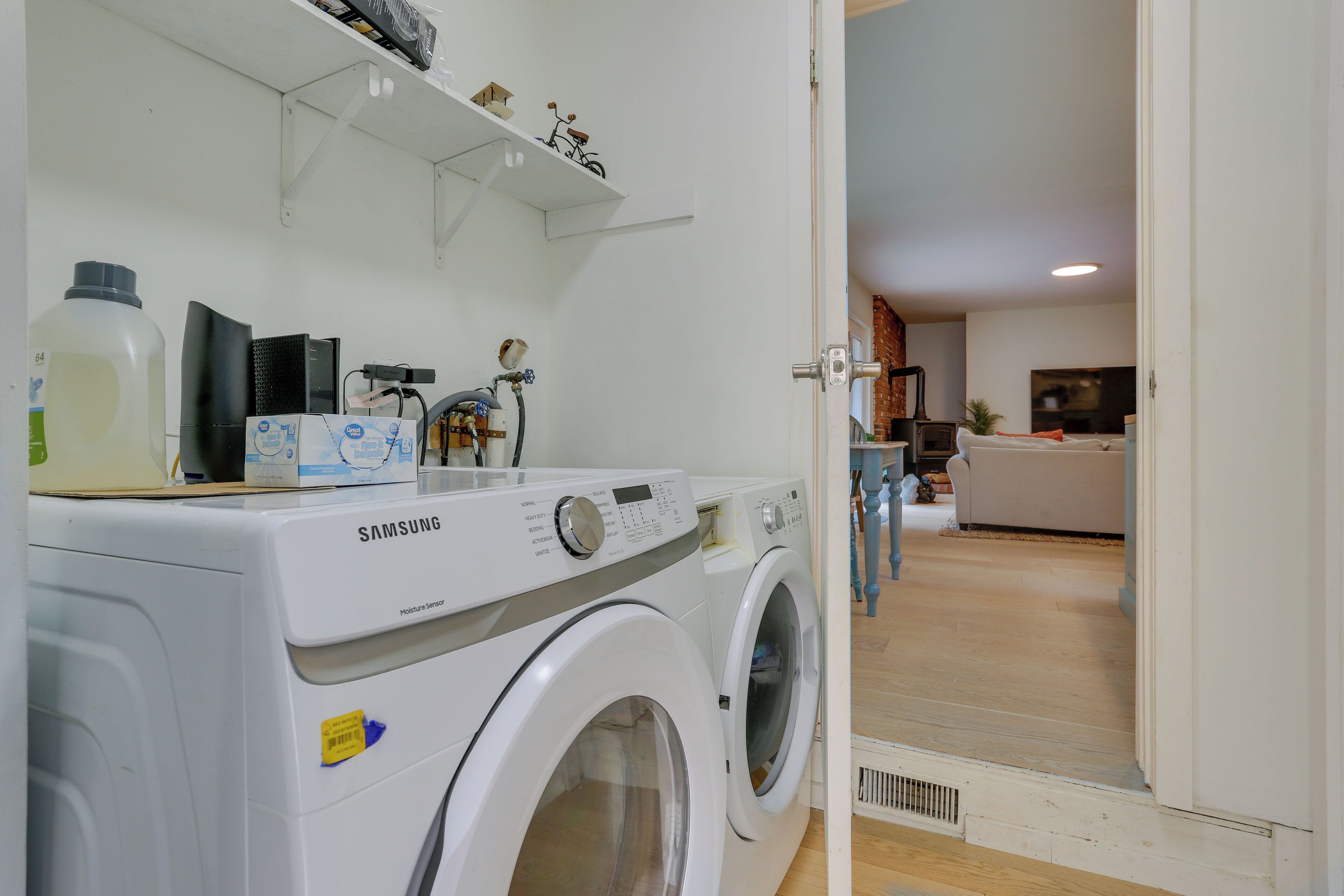 Un área de lavandería con una lavadora Samsung, un estante con objetos pequeños y vista a una sala de estar con sofá y televisor.