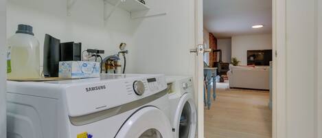 Un área de lavandería con una lavadora Samsung, un estante con objetos pequeños y vista a una sala de estar con sofá y televisor.