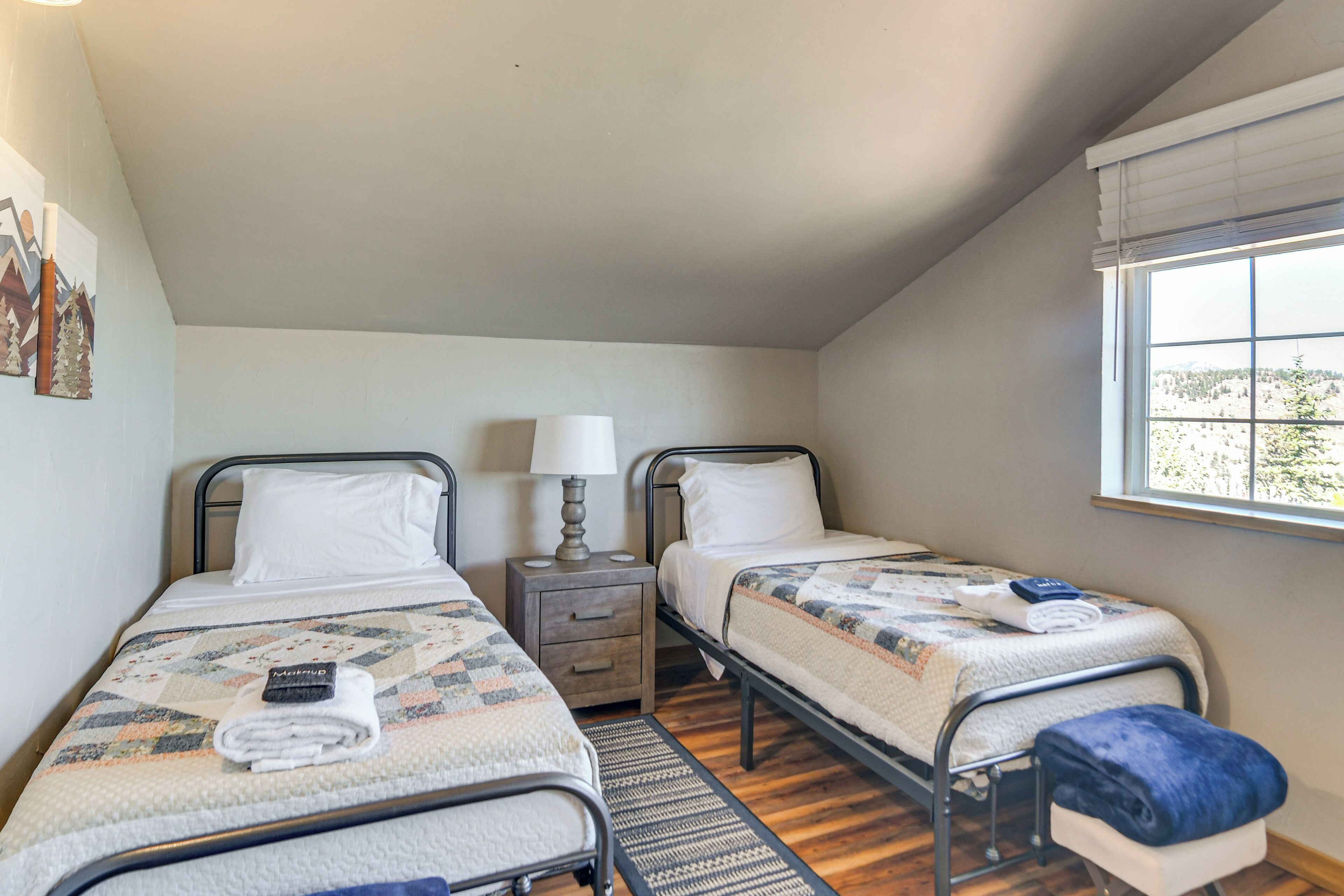 A bedroom with two single beds, a nightstand with a lamp, a window with blinds, and a view of trees outside.