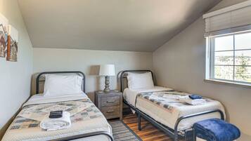 A bedroom with two single beds, a nightstand with a lamp, a window with blinds, and a view of trees outside.