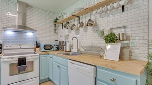 Una cocina con paredes de azulejos blancos, encimera de madera, extractor de acero inoxidable, lavavajillas y un fregadero.