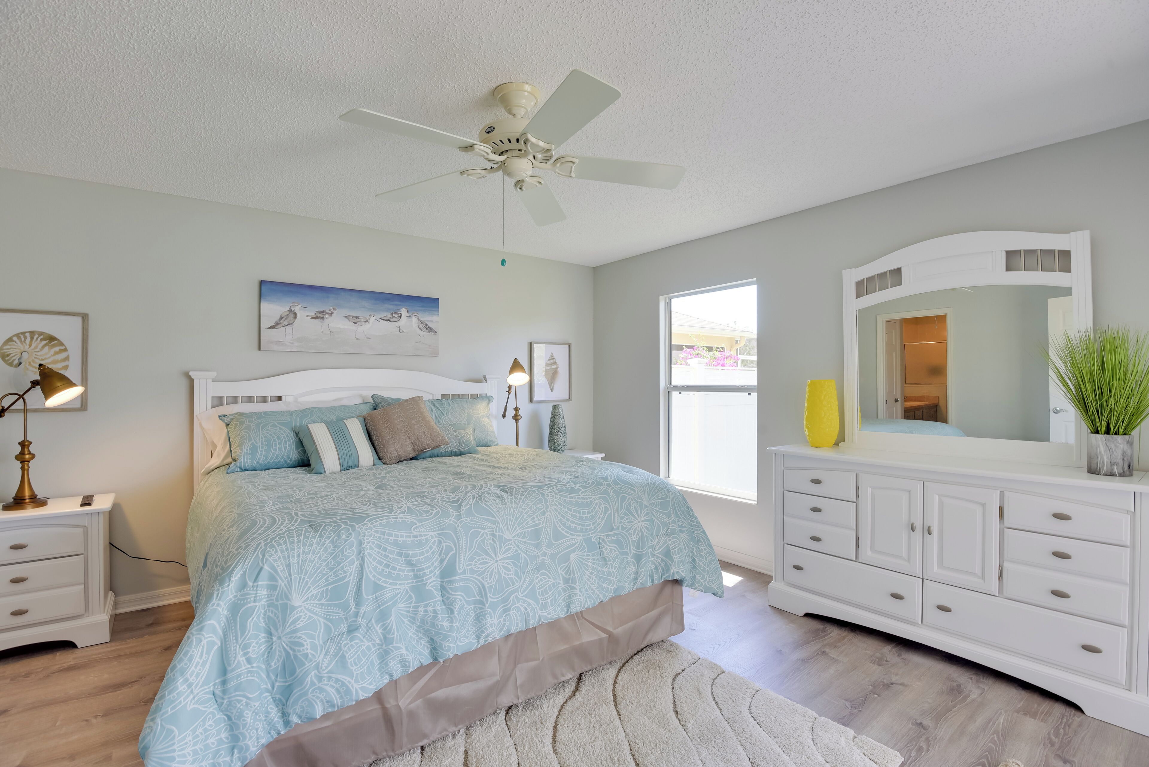 A bedroom with a bed, nightstands, a dresser, a ceiling fan, a large window, and artwork on the walls.