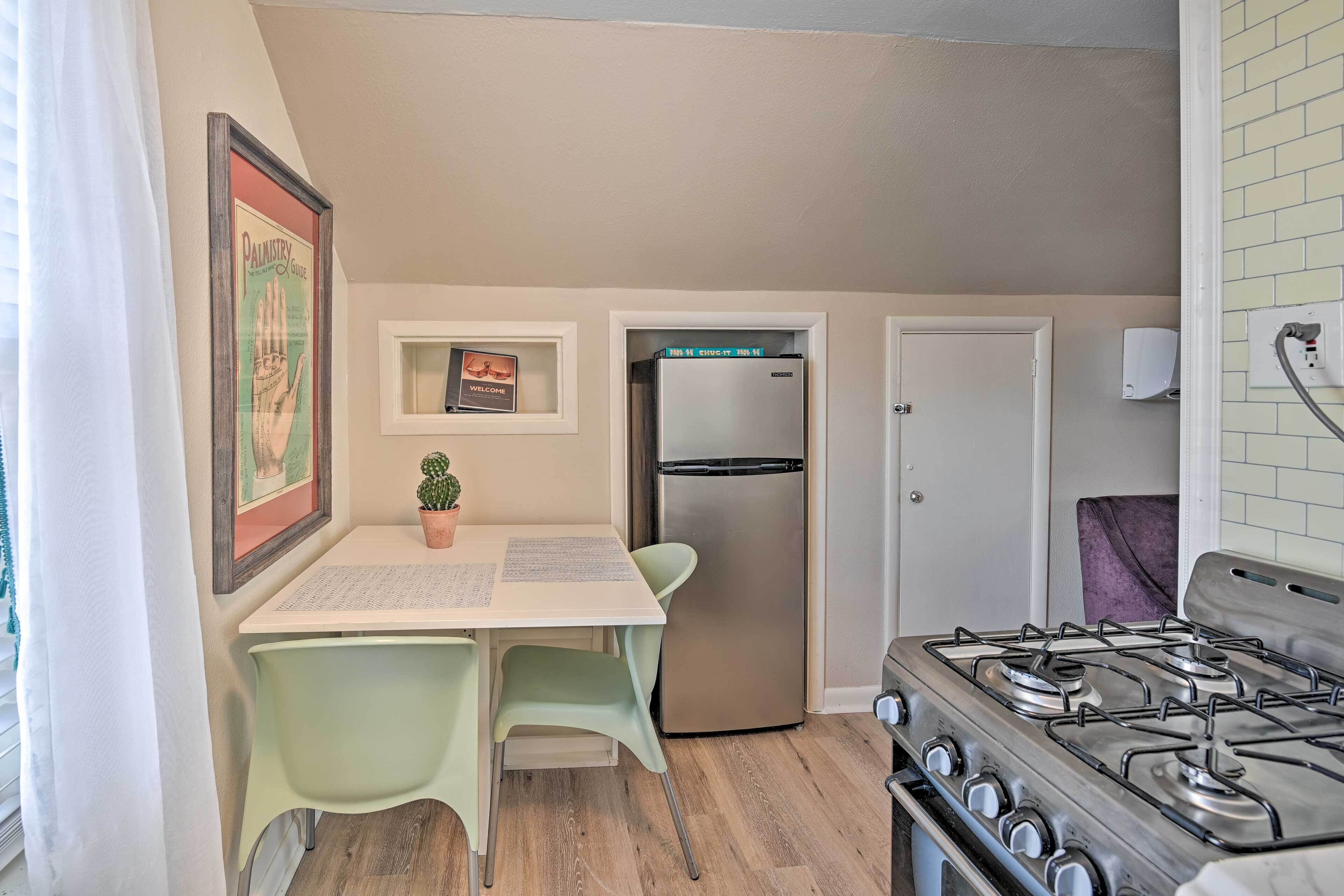 A compact kitchen with a gas stove, a refrigerator, a dining table with chairs, a small potted plant, and a framed picture on the wall.
