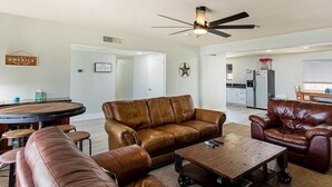 A spacious living room with a brown leather sofa, a coffee table, and a dining area with a round table. A ceiling fan is visible, and there is a kitchen in the background. - Havasu Warbler - Hot Tub, BBQ, Parking, FirePit (Lake Havasu City)