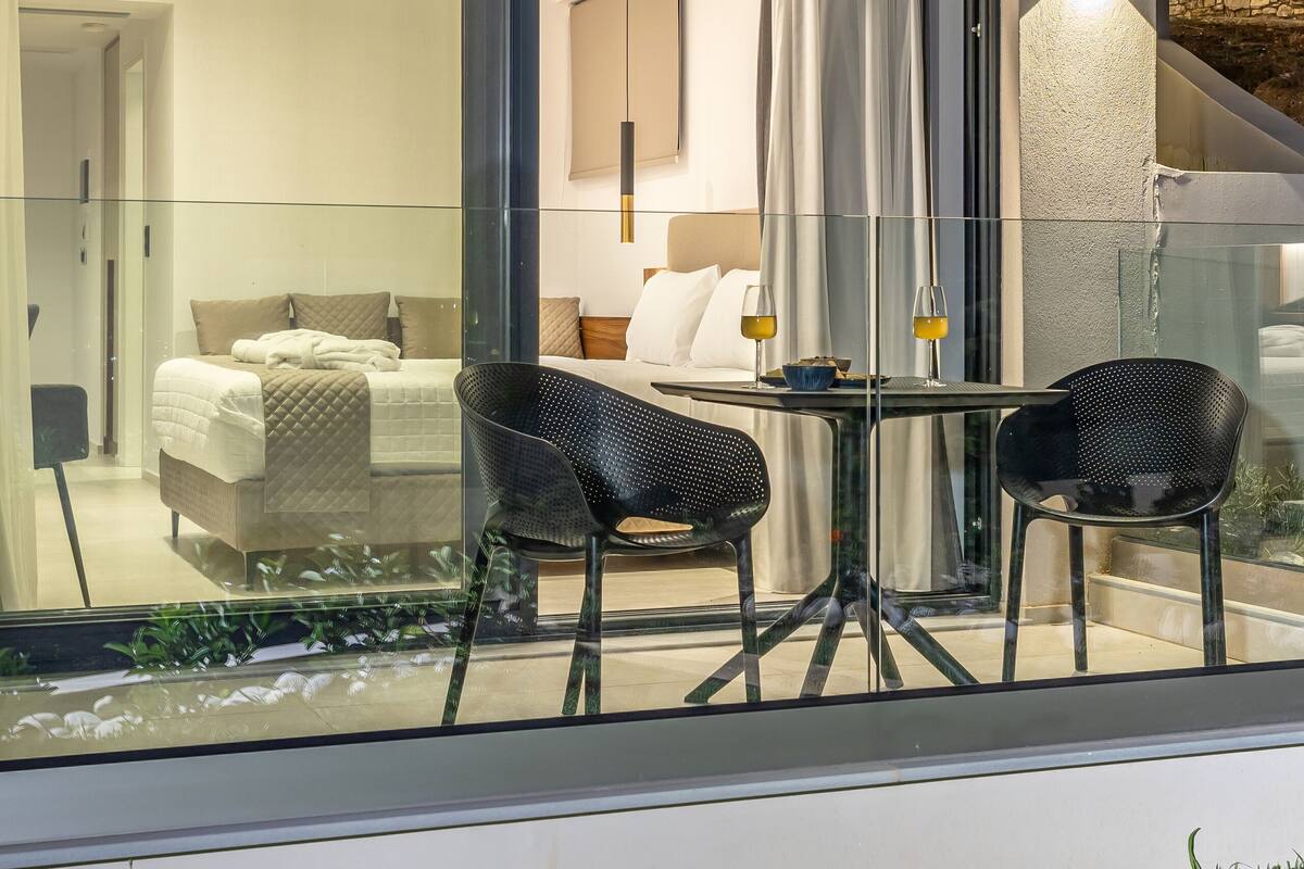 A modern hotel room with a glass partition, a bed, a dining area with two chairs, and a large window with curtains.