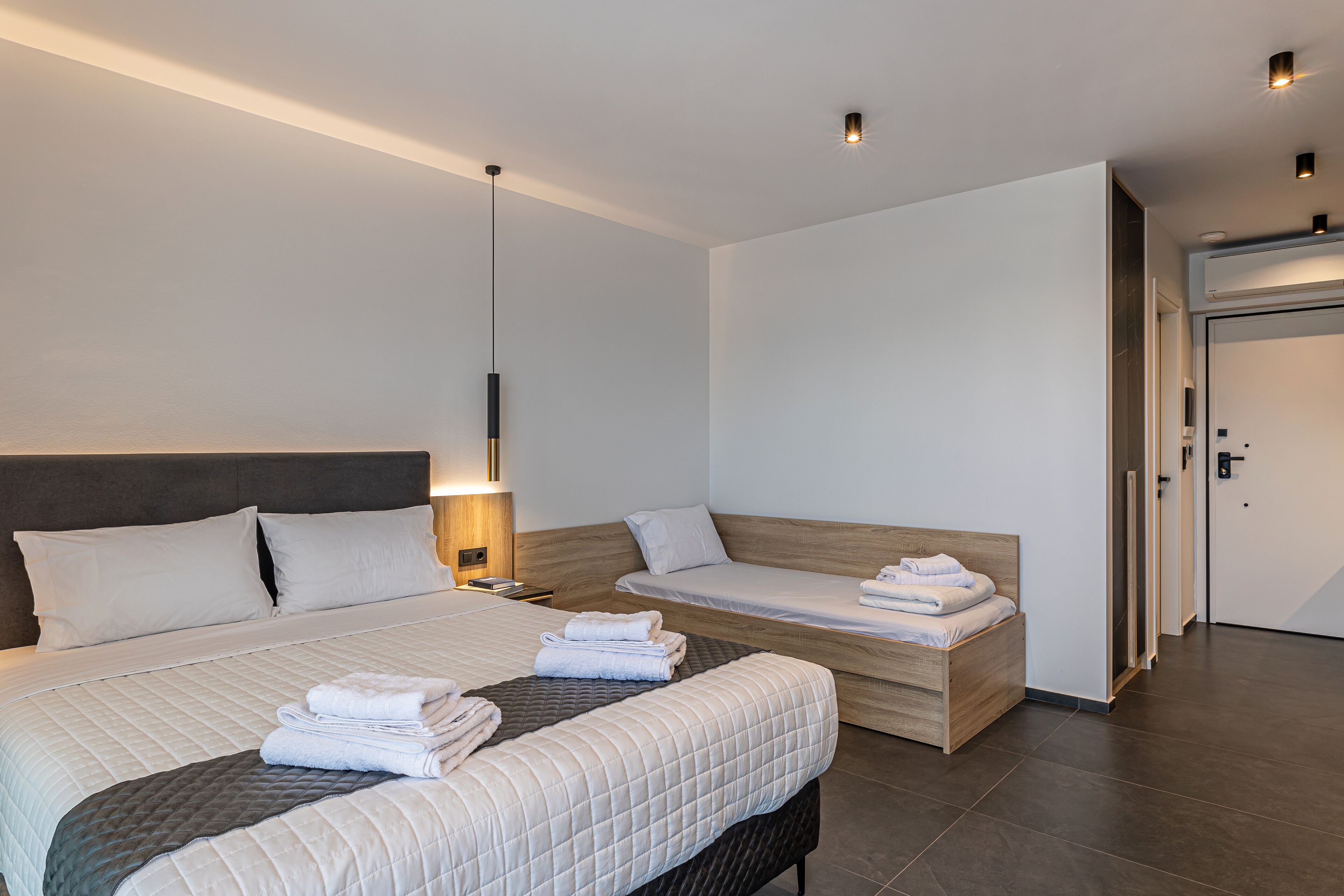 Una habitación de hotel moderna con una cama grande, una cama más pequeña, una mesita de noche, una lámpara y una puerta.
