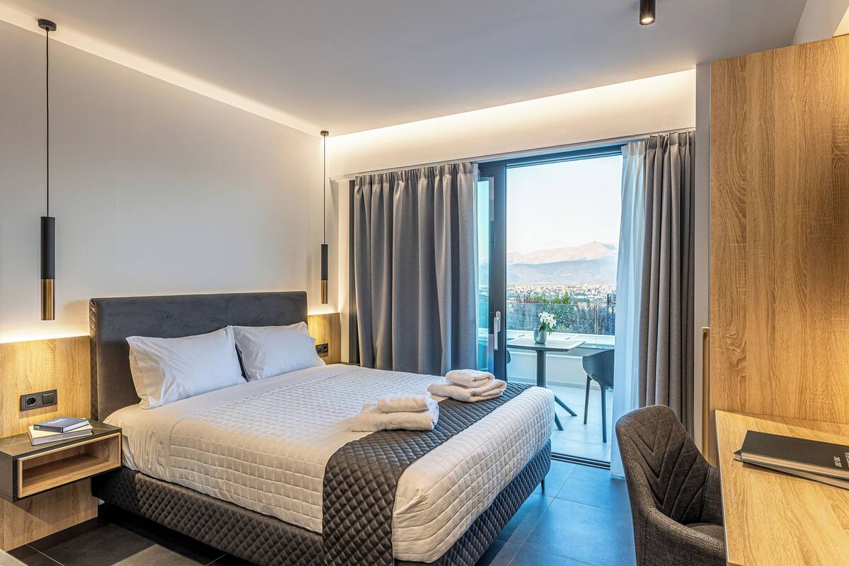 A modern hotel room with a large bed, a desk, and a view of the city through the window.