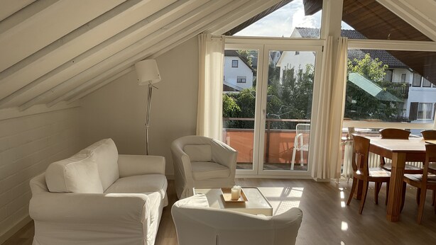 Un vaste espace de vie doté d’un haut plafond, d’un canapé blanc, d’un fauteuil, d’une table à manger avec des chaises et d’une grande porte-fenêtre coulissante donnant sur un patio extérieur.
