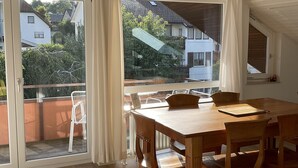 Une pièce spacieuse avec une table et des chaises en bois, de grandes fenêtres donnant sur le quartier et un plafond avec des poutres apparentes.