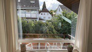 Une pièce avec une table en bois et deux chaises, une fenêtre donnant sur des maisons et des arbres, et une chaise blanche sur laquelle est posé un objet décoratif sur le rebord de la fenêtre.
