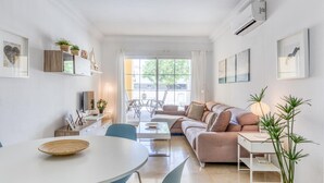 Een moderne woonkamer met een bank, eettafel en stoelen. Een groot raam zorgt voor daglicht en uitzicht op de omgeving.