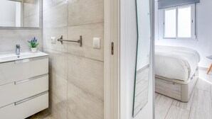 Une salle de bain moderne avec un lavabo blanc, un miroir et un porte-serviettes. On peut apercevoir un lit avec une literie blanche dans la pièce adjacente.