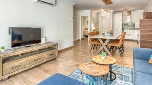 A modern living room with a TV, wooden furniture, a dining table with chairs, and a kitchen area in the background. - Modern apartment in Estepona, ground floor with terrace and pool, close to port and beach (Estepona)