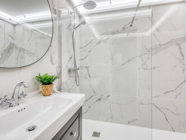 Un baño con lavamanos de mármol blanco, un espejo redondo, ducha con cabezal de ducha de mano y una planta en maceta.