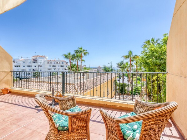 Un balcón con mobiliario de mimbre y vistas a una zona costera con edificios y palmeras.