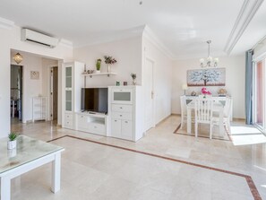 Uma sala de estar moderna com uma TV de tela plana, armários brancos, uma mesa de jantar com cadeiras e uma janela grande.