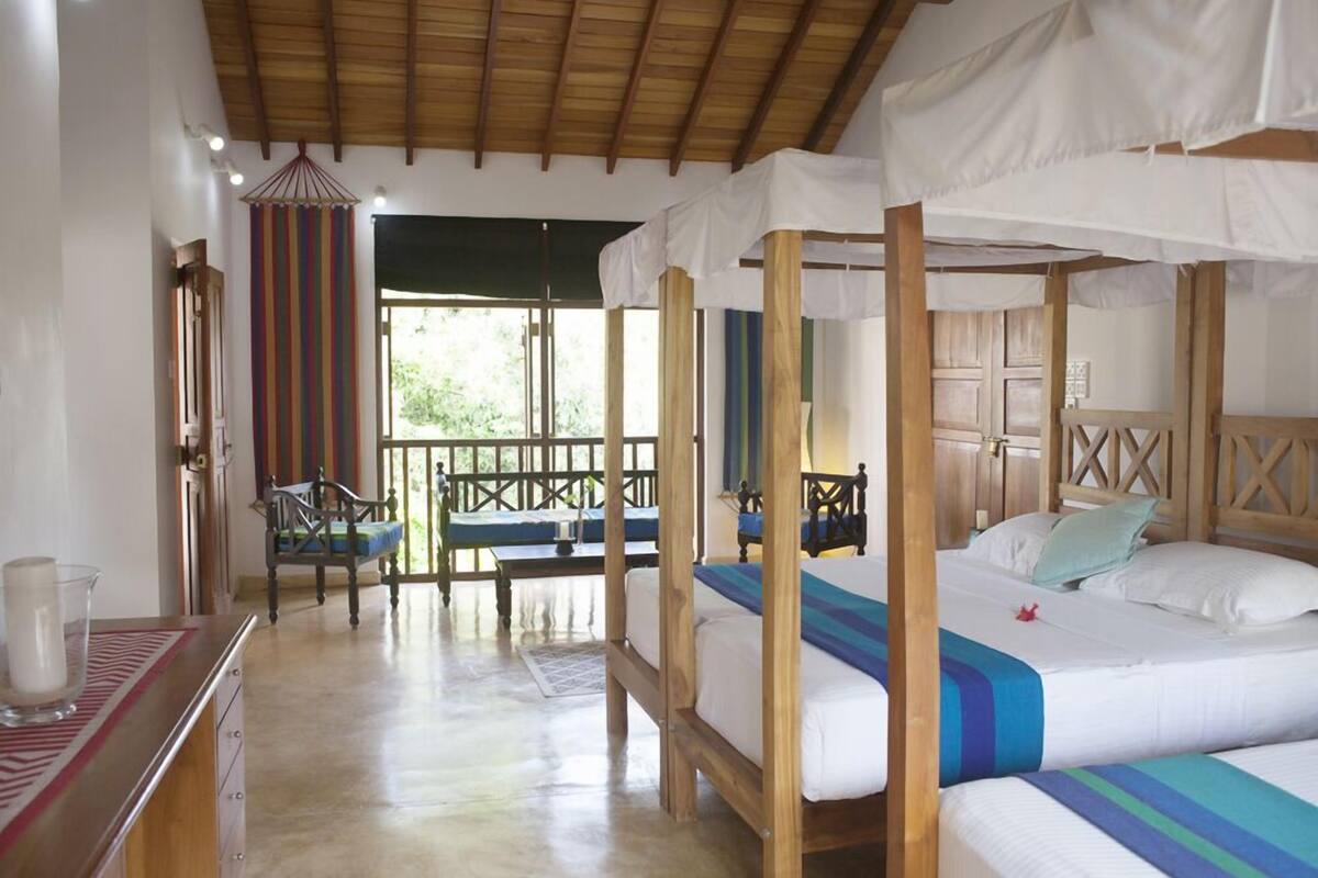 A room with a wooden four-poster bed, a small table, and a chair. There is a window with a view of greenery outside.