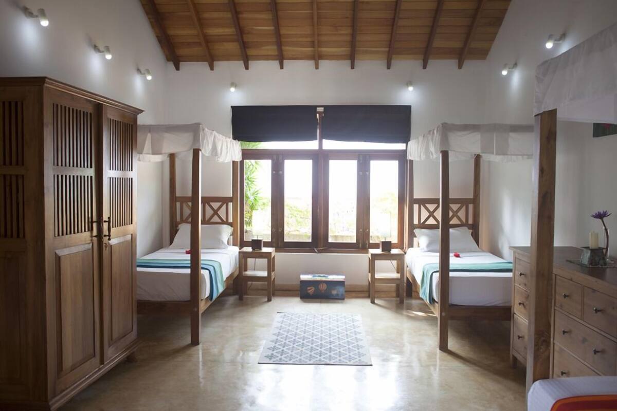 A room with two beds, a wardrobe, a window with curtains, and a ceiling with wooden beams.