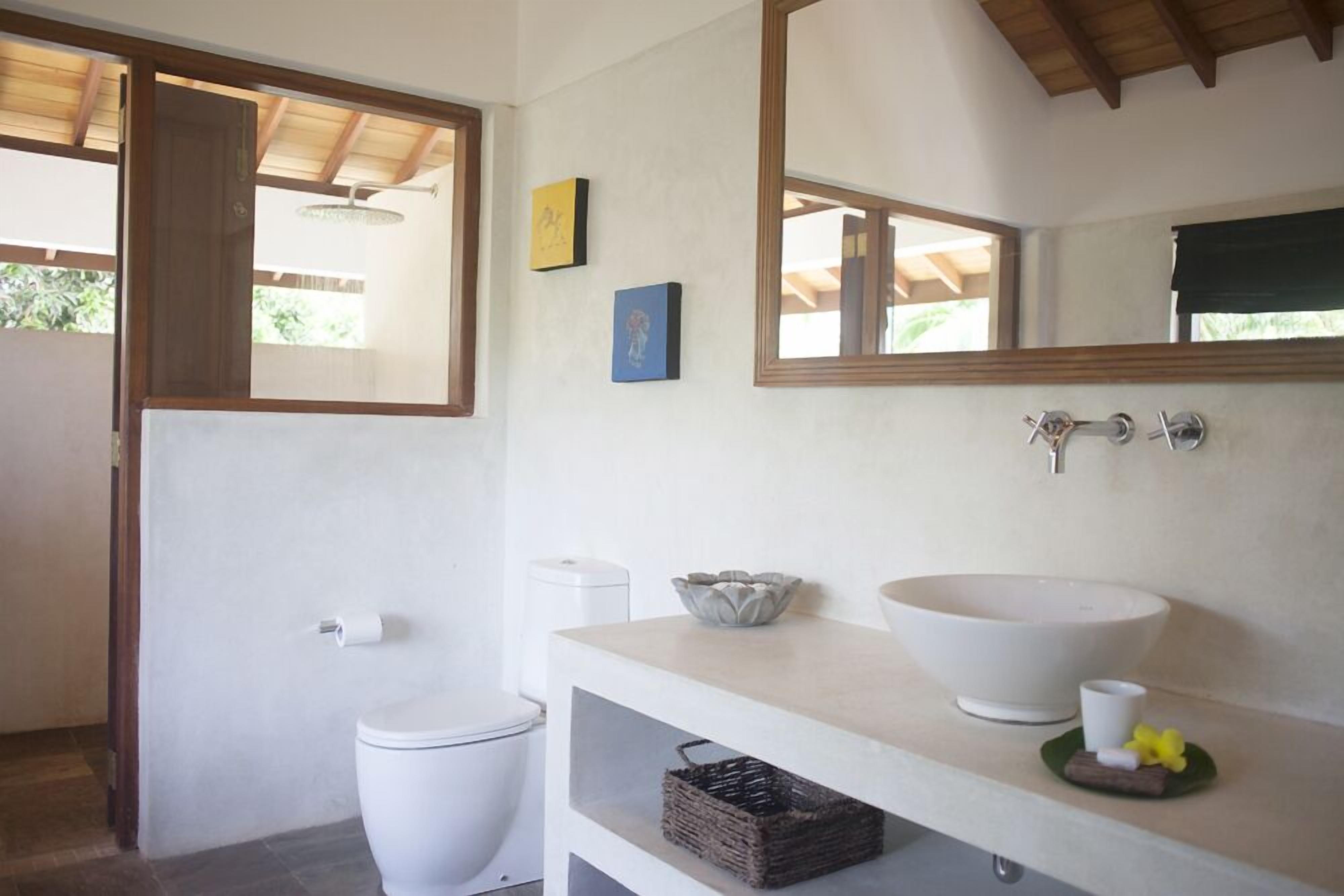 Une salle de bain comprenant des toilettes, un lavabo avec un grand bac, un miroir et une petite étagère avec un panier.