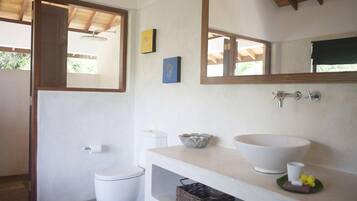 Une salle de bain comprenant des toilettes, un lavabo avec un grand bac, un miroir et une petite étagère avec un panier.