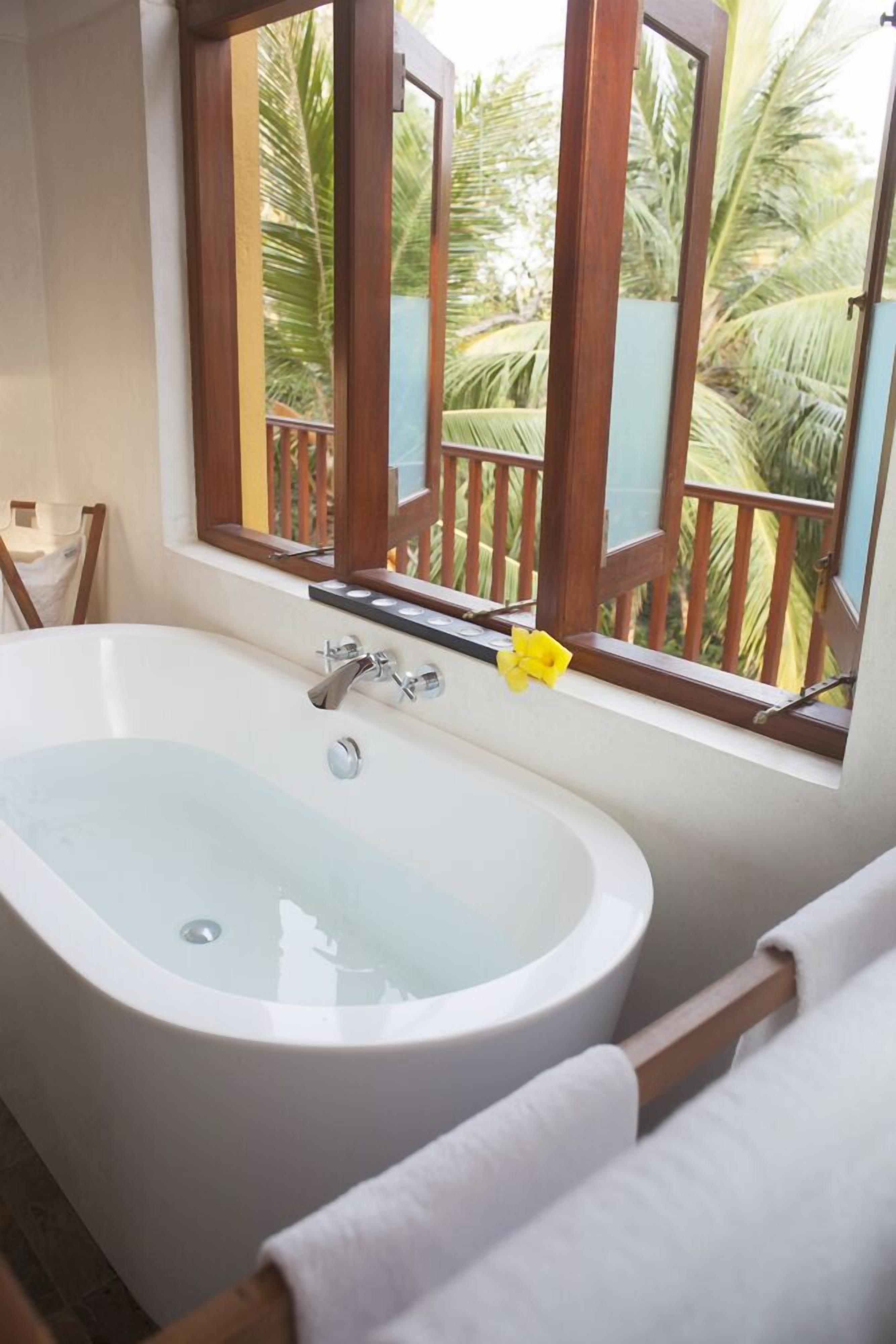 Un baño con una bañera independiente, una ventana que ofrece vistas a palmeras y una barandilla de madera.