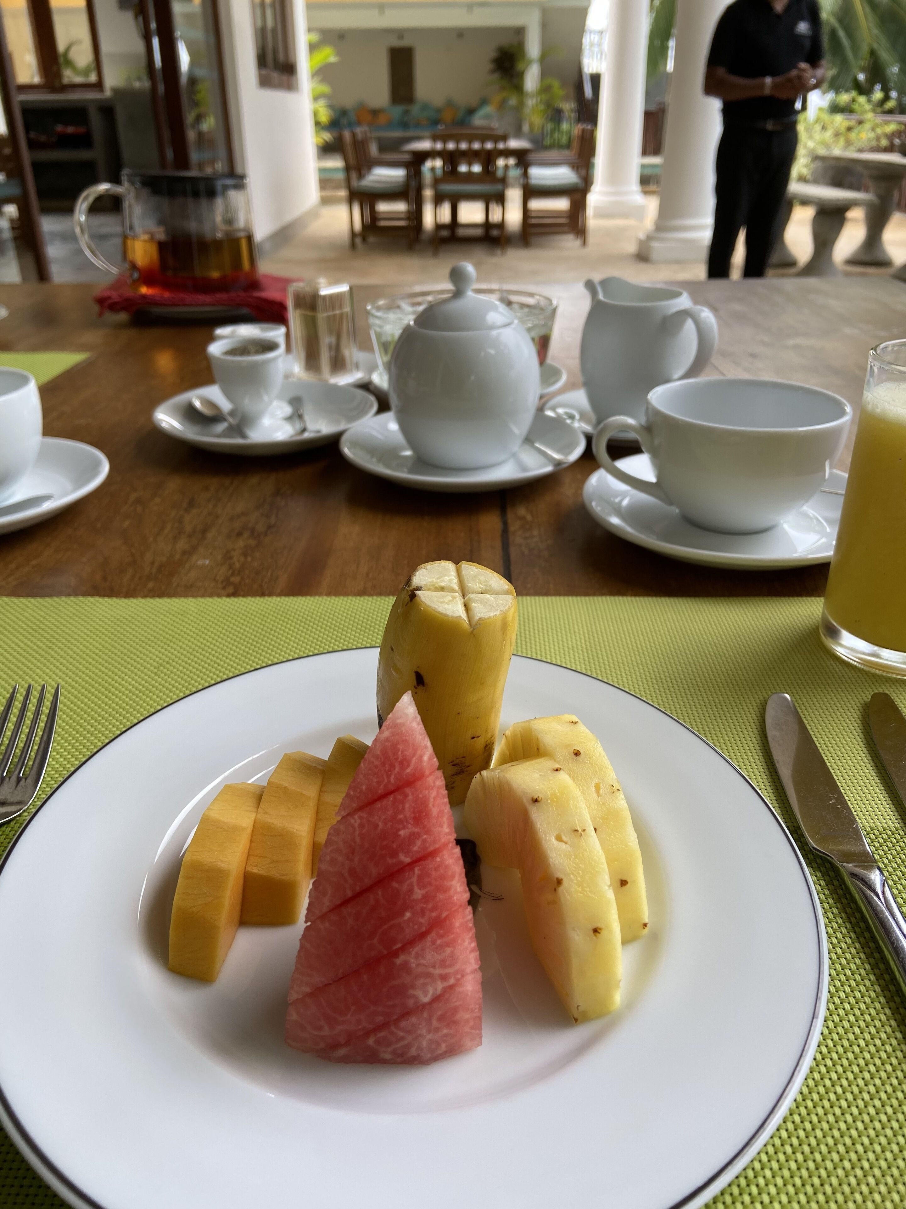 Una mesa de comedor con un plato de fruta, una tetera, tazas y un vaso de jugo.