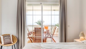 A hotel room with a bed, a wicker chair, a nightstand with a lamp, and a view of outdoor seating through a large window. - 3-bedroom duplex with communal pools and Wi-Fi in Estepona (Estepona)