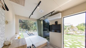 A modern bedroom with a bed, a desk, a chair, a TV, and a large sliding glass door to a garden.