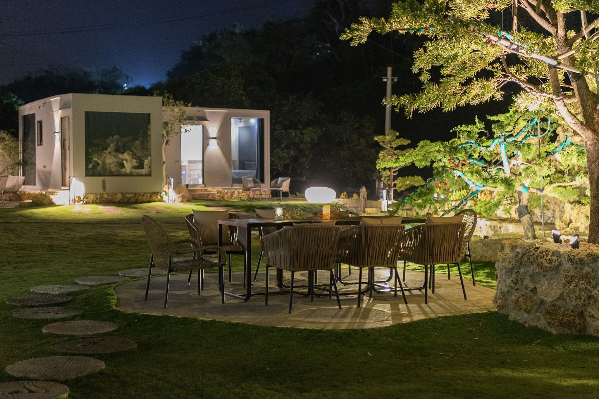A modern outdoor dining area with a table and chairs, a small building with a glass door, and a tree with hanging lights.