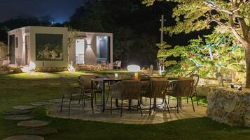 A modern outdoor dining area with a table and chairs, a small building with a glass door, and a tree with hanging lights.