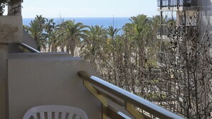 A balcony with a view of the sea, palm trees, and a building with balconies.