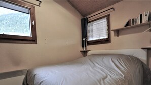 A bedroom with a bed, a window, a shelf with books, and a view of a forest.