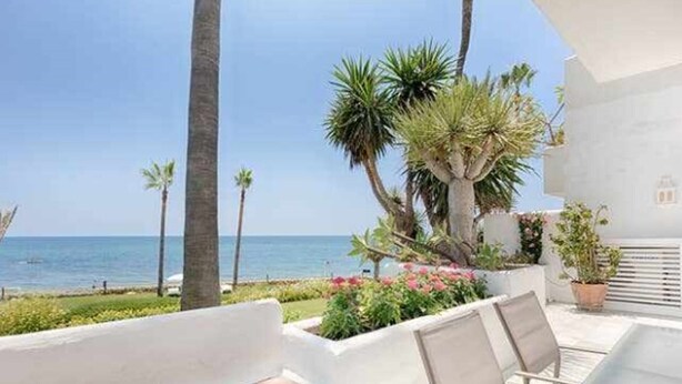 A balcony with a view of the sea, palm trees, and a white building. - Luxury beachfront apartment with pool, gardens, tennis courts and restaurant in Estepona (Estepona)