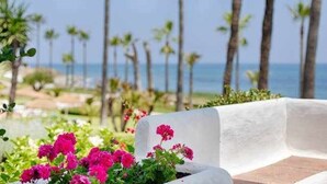 A white bench with pink flowers in front of a beach with palm trees and the ocean in the background. - Luxury beachfront apartment with pool, gardens, tennis courts and restaurant in Estepona (Estepona)