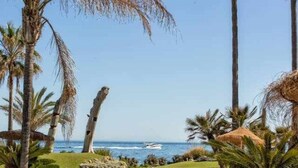 A pool area with palm trees, a view of the ocean, and a thatched structure. - Luxury beachfront apartment with pool, gardens, tennis courts and restaurant in Estepona (Estepona)