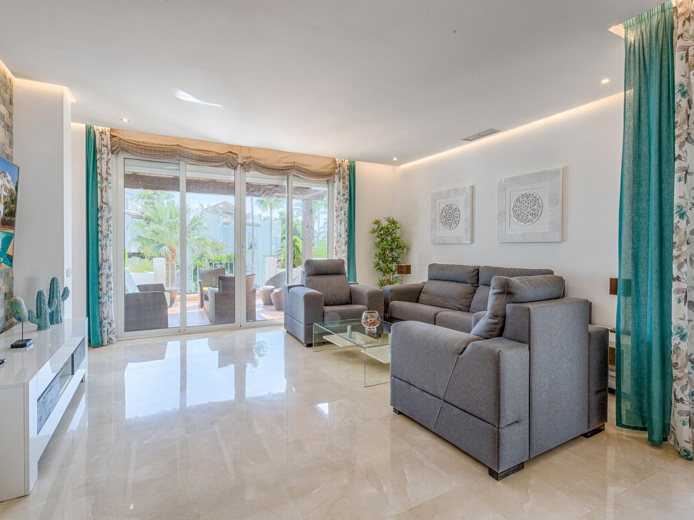 Una sala moderna con un sofá gris, una mesa de centro de vidrio y vistas a un patio con mobiliario exterior.