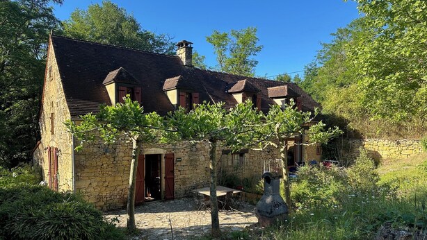 Una casa de piedra con techo inclinado y contraventanas de madera, rodeada de follaje y un sendero empedrado.