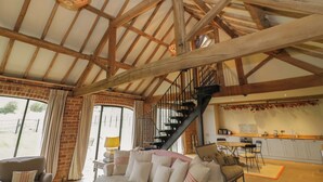 A spacious living area with a high ceiling, exposed wooden beams, a staircase, and a kitchen area with modern appliances. - The Carthouse (Ledbury)