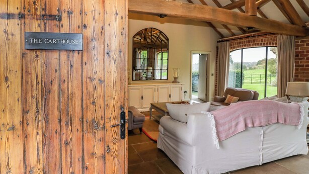 A rustic interior with a wooden door labeled "THE CARTHOUSE," a white sofa, and a large window offering a view of the outdoors. - The Carthouse (Ledbury)