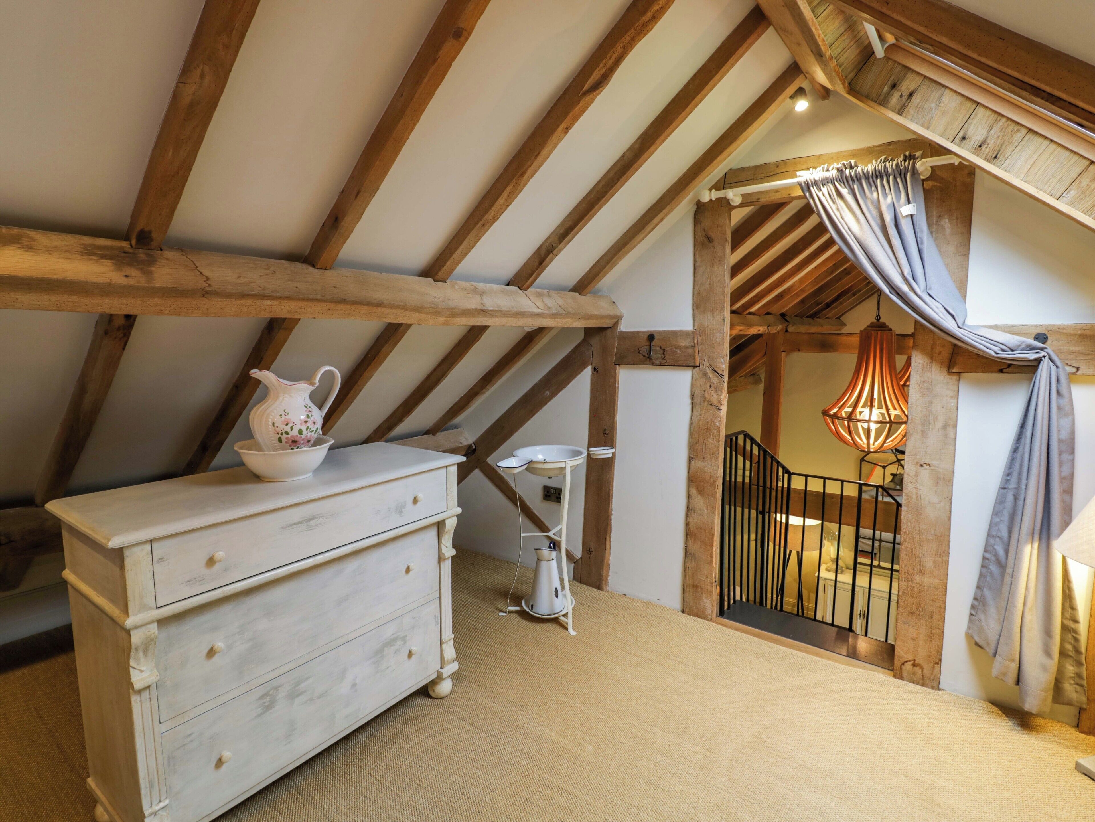 Un dormitorio rústico en el ático con vigas de madera a la vista, un aparador blanco, una mesita con una jarra y una escalera que lleva a un balcón.