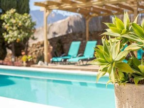 A poolside area with a concrete planter holding green plants, a wooden pergola, and a mountain backdrop.