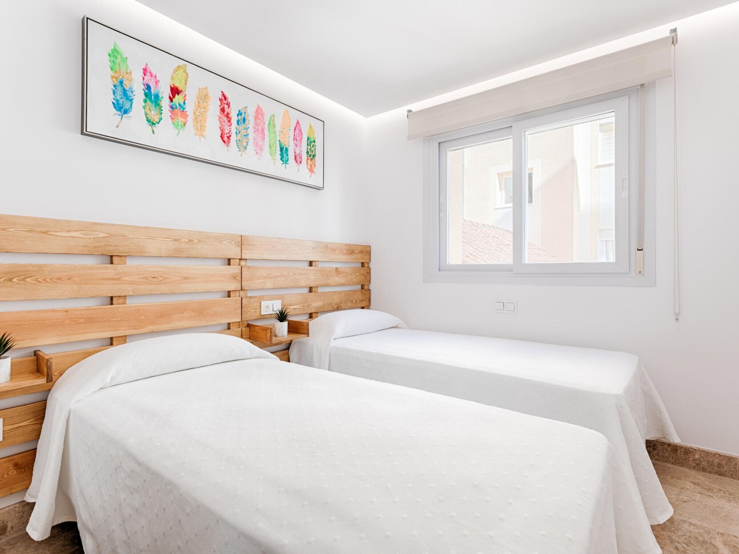 A hotel room with two beds, a wooden headboard, a window, and a colorful feather artwork on the wall.
