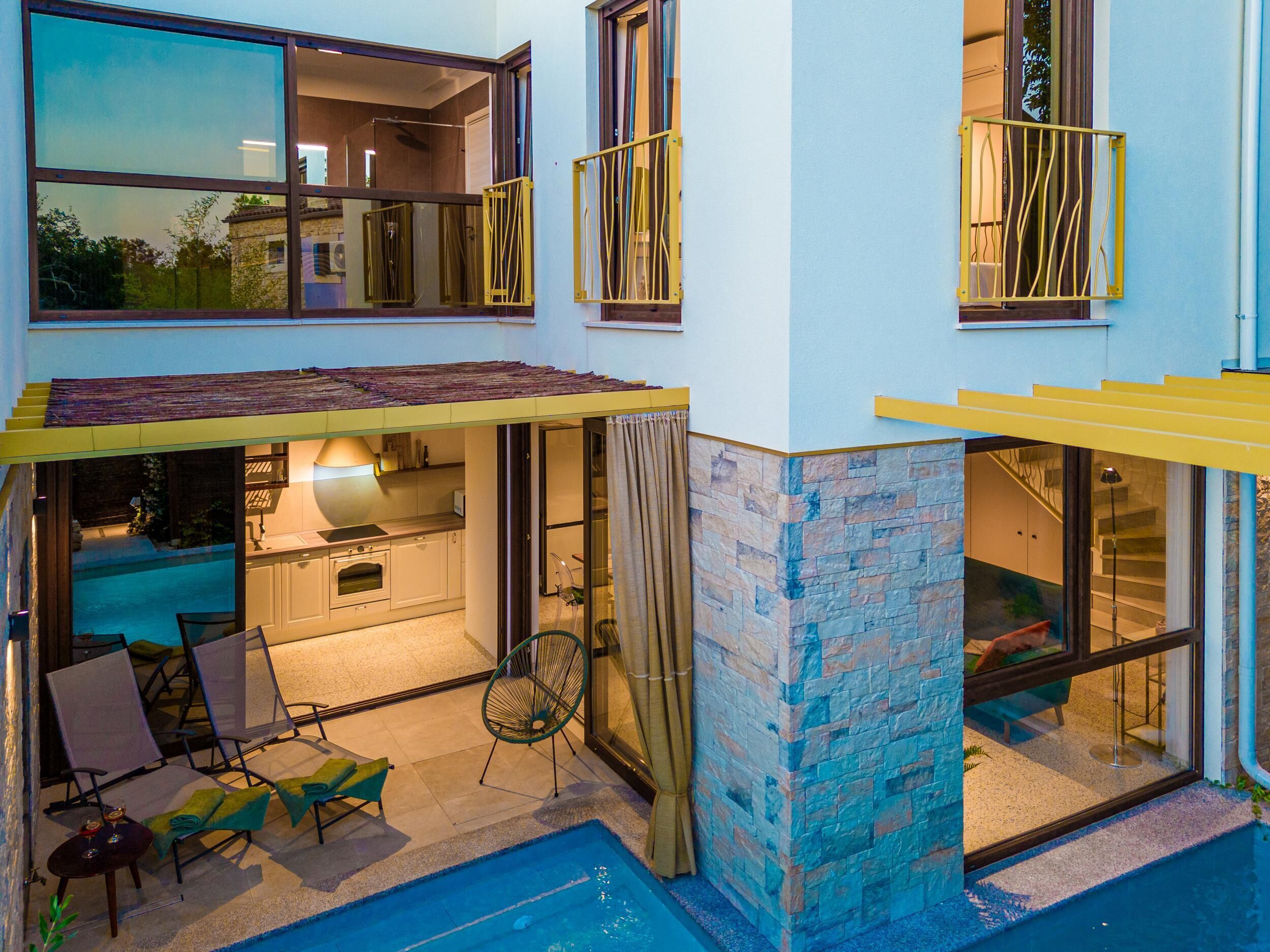 A two-story house with a swimming pool, outdoor seating, and a kitchen visible through glass doors.