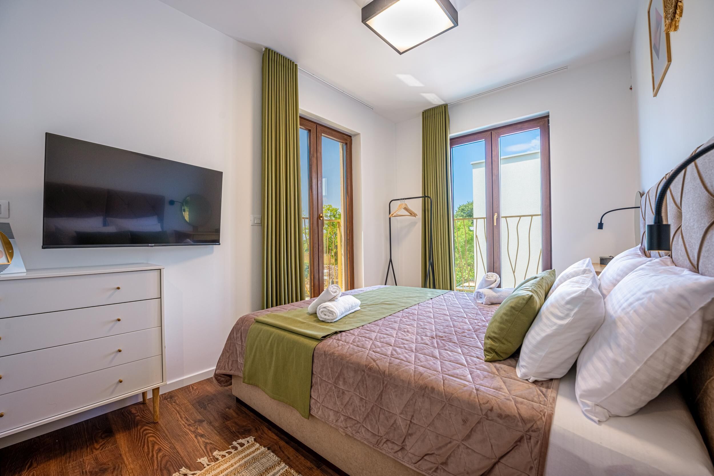 Une chambre à coucher avec un lit, un téléviseur fixé au mur, une commode et une fenêtre avec des rideaux.
