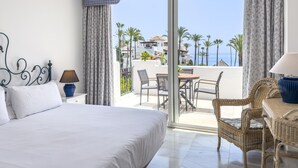 A hotel room with a bed, a small table with chairs, a wicker chair, a lamp, and a view of the sea and palm trees.