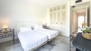 A double bed with white linens, a small bedside table with a lamp, a large wardrobe with louvered doors, and a wicker table with a basket.